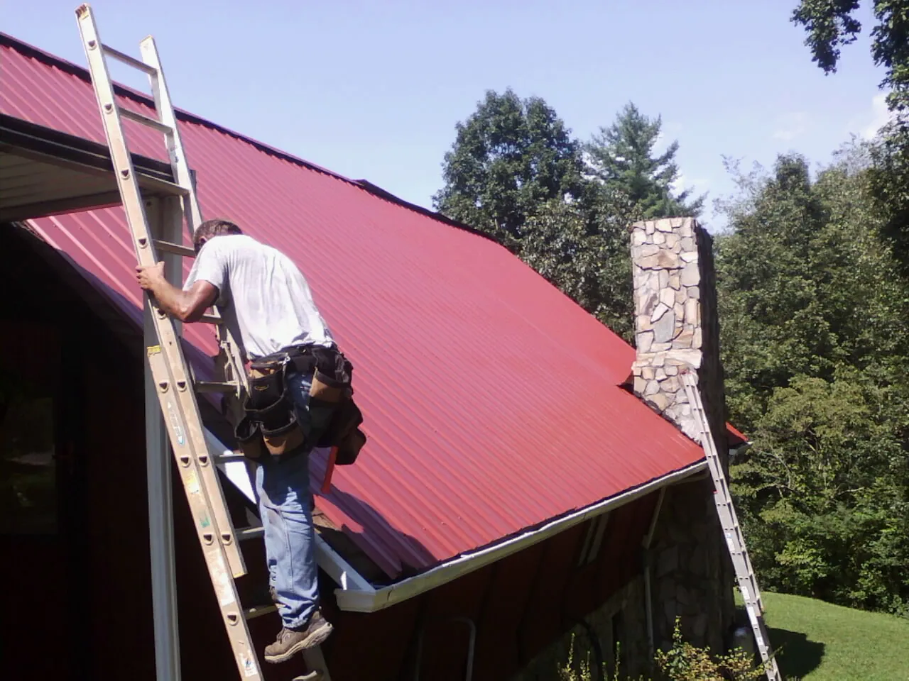 Roofer on a ladder beside a red metal roof and stone chimney — Soffit & Fascia Repair by Roofing Brothers in Beech Bluff